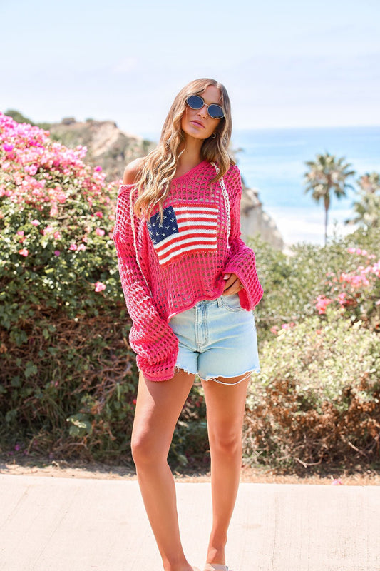 Woman in sunglasses and the All American Open Knit Hoodie stands outdoors near flowers and the beach on a sunny day.