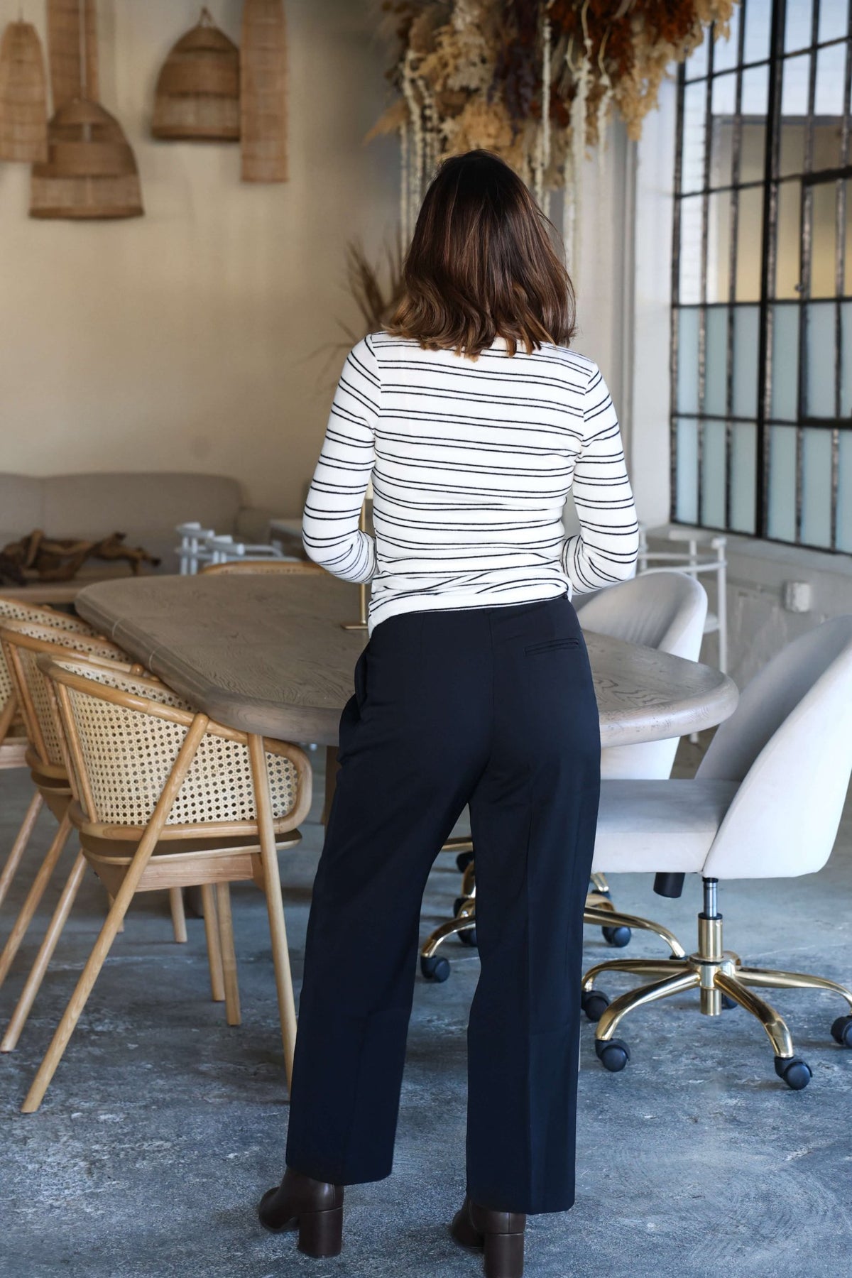A woman in a striped top and Anabelle Black Pleated Trousers stands at a table in a modern dining area.