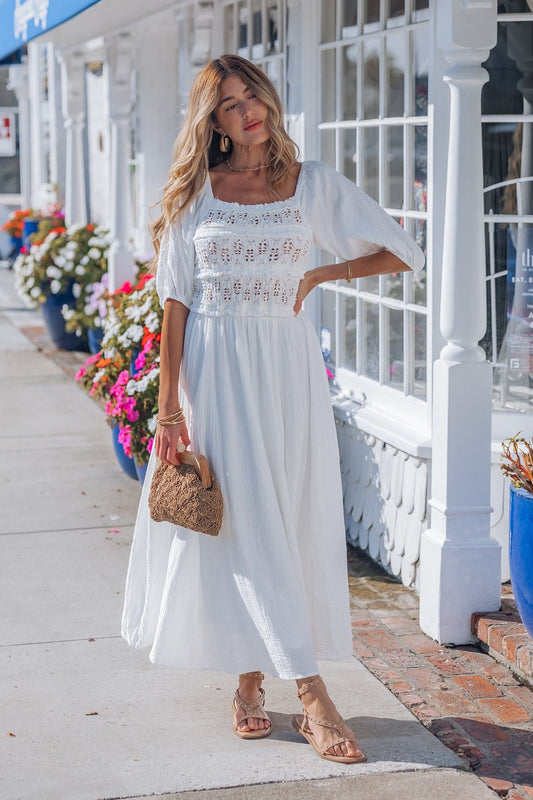Woman wears the Angelica White Ruched Midi Dress - FINAL SALE, holding a woven bag on a sunny, flower-lined sidewalk.
