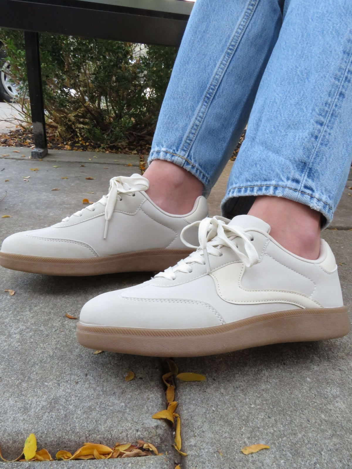 Wearing beige Remus Lace Up Sneakers, someone in light blue jeans stands on leaf-strewn stone pavement.