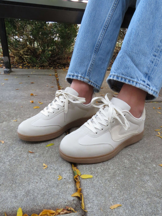 Person wearing Beige Remus Lace Up Sneakers - FINAL SALE and blue jeans sits on a bench outdoors among fallen leaves.