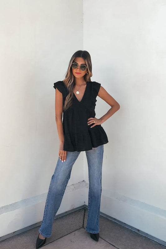 Woman wearing the Black Ruffle Sleeve Textured Top- FINAL SALE, blue jeans, and black heels stands confidently by a white wall.