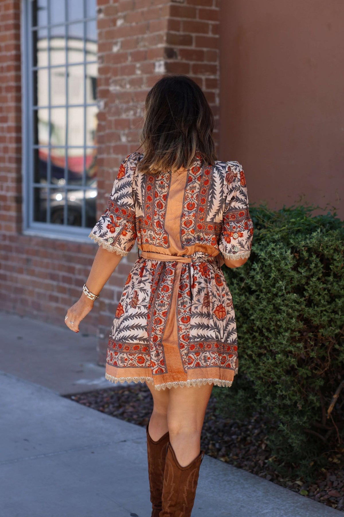 Woman in a Botanical Rust Print Belted Mini Dress and brown boots walks on a sidewalk by a brick building and green bush.