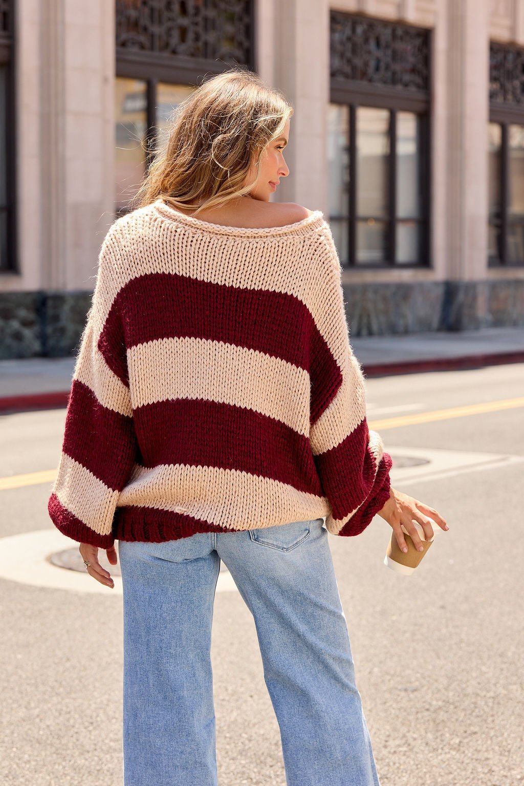 Woman in a Camel & Wine Striped Sweater with balloon sleeves holds a coffee cup, facing away from the camera.