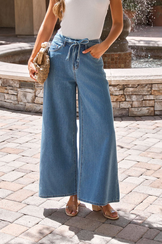 Woman in Denim Belted Wide Leg Pants-FINAL SALE, white sleeveless top, holding a woven clutch by a sunny stone fountain.
