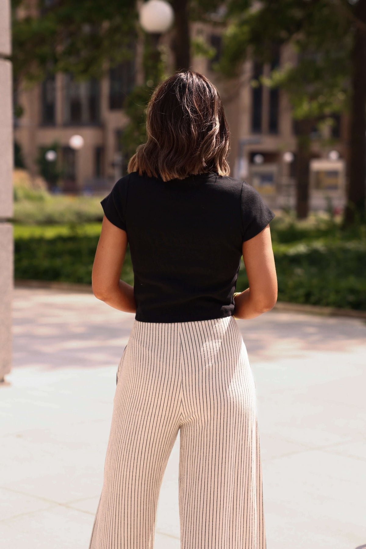 A woman wears the Everyday Essential Black Tee-FINAL SALE with striped pants outdoors. The 100% cotton top ensures a comfy fit.