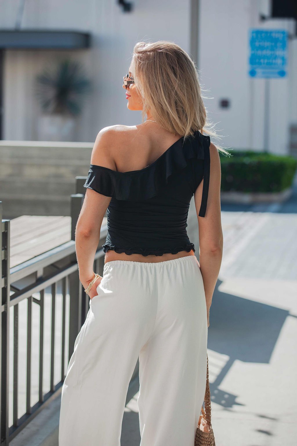 A woman wears a black Free People Isla Off The Shoulder Top - Black-FINAL SALE and white pants, outdoors with an asymmetrical ruffled neckline.