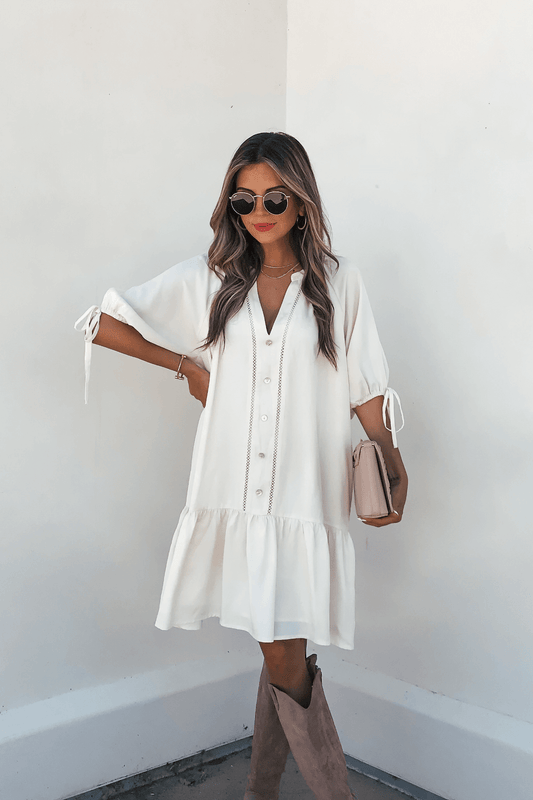 Woman in the Laurie Cream Button Down Mini Dress holds a beige clutch, styled with brown boots and sunglasses against a white wall.