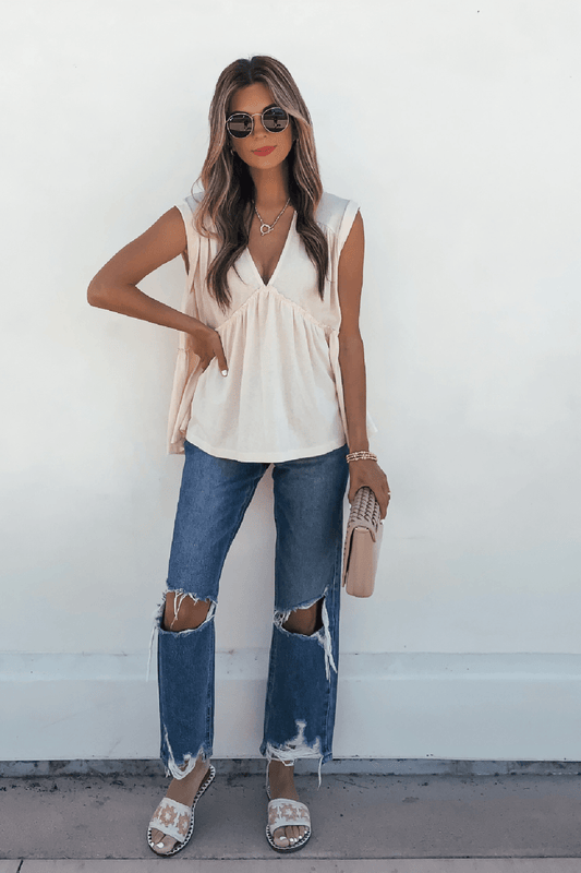Woman in sunglasses and Lena’s Carefree Afternoon Cream Babydoll Top poses stylishly against a white wall in jeans, clutch, and sandals.