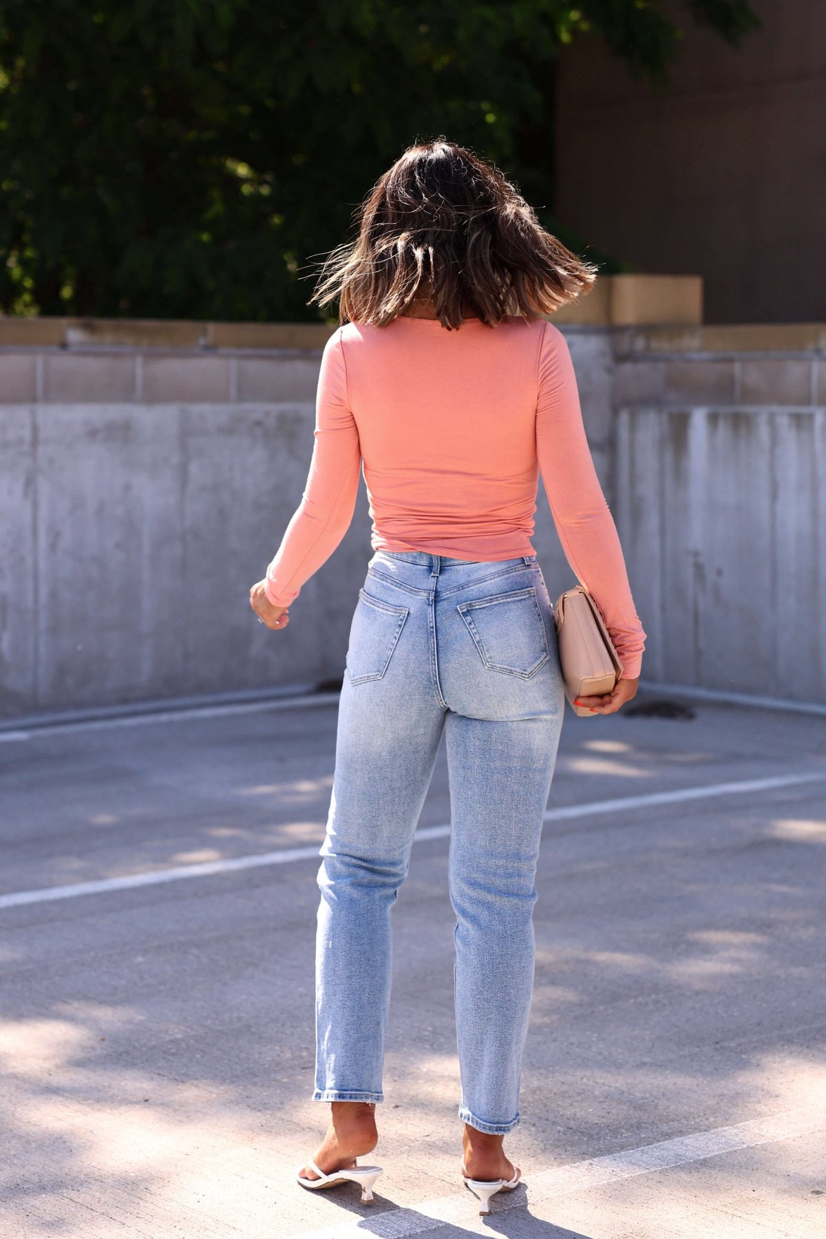 A woman outdoors, facing away, wears Light Wash High Rise Cropped Straight Jeans - FINAL SALE with a pink top, sandals, and beige clutch.