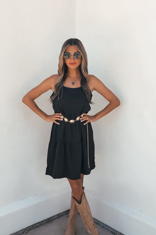 Woman wears the On Point Tiered Mini Dress - Black, sunglasses, brown boots, and a gold-buckled belt against a white wall.