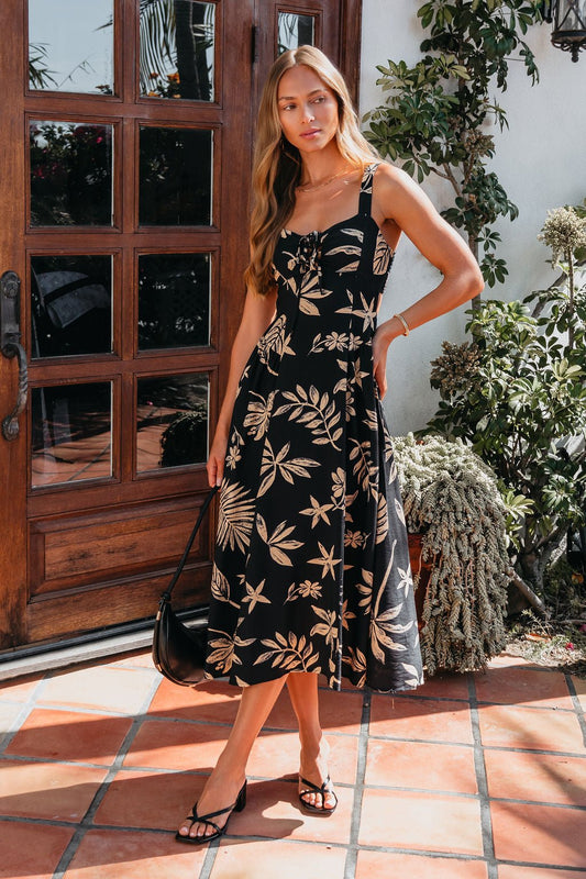 Woman in a Paloma Black Tropical Print Midi Dress stands by a wooden door with a black bag, surrounded by lush plants.