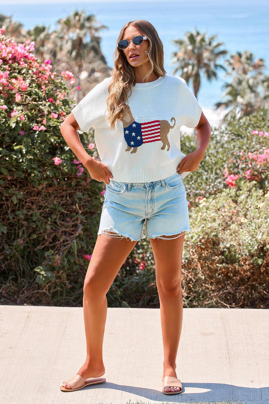 Woman in a Patriotic Pup Knit Sweater—Cream, denim shorts, and sandals stands outdoors by flowers and palm trees.