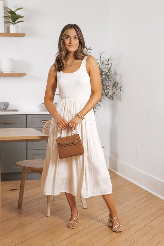 Woman in the Scoop Neck Pleated Skirt Midi Tank Dress holds a tan bag, standing in a minimalistic room with light wood floors.