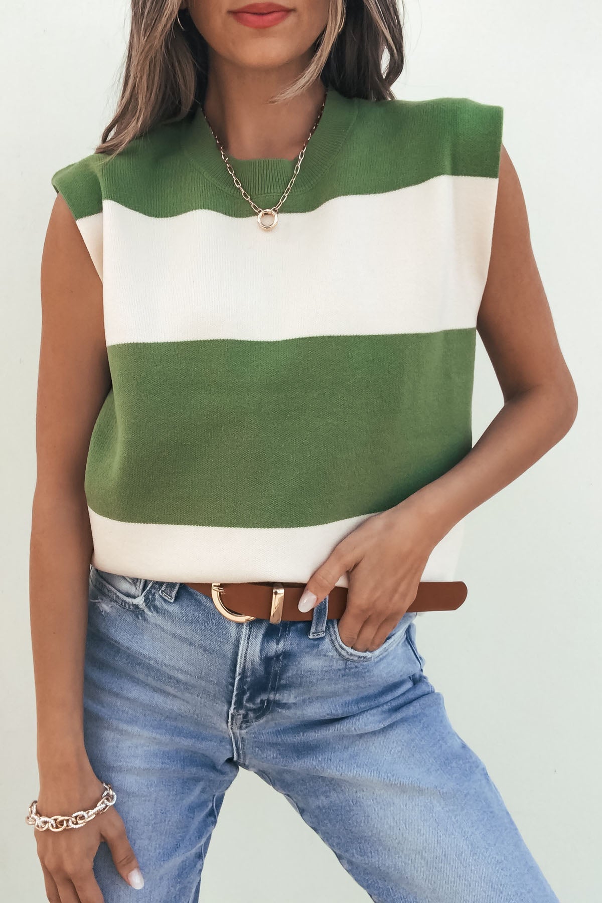 Woman wears a Sleeveless Green and Cream Striped Sweater-FINAL SALE, blue jeans, brown belt, gold necklace, and silver bracelet.