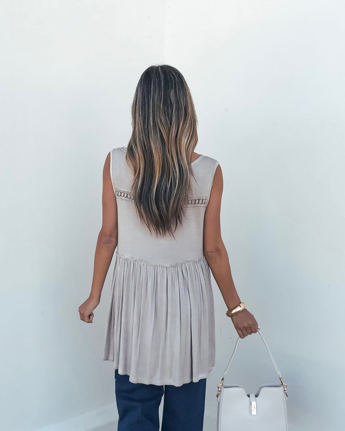 Woman with long wavy hair faces away in Taupe V Neck Flowy Top-FINAL SALE, dark pants, holding white bag against a light background.