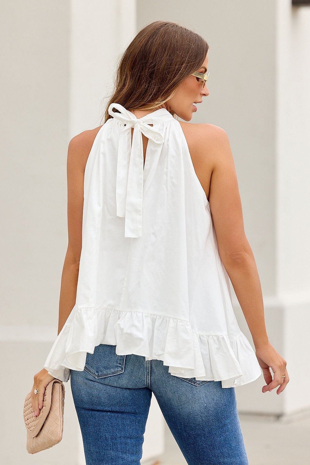 Woman in The Harlow Ruffle Top - White, blue jeans, and holding a beige clutch, seen from behind.