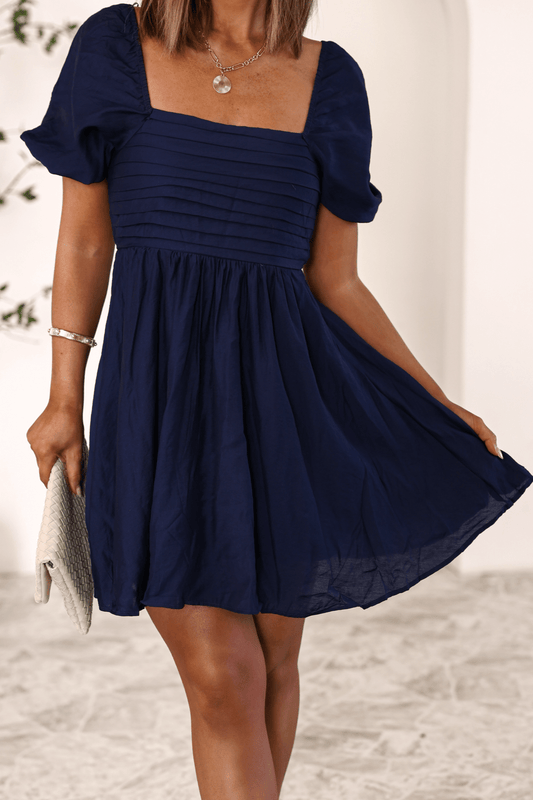 Woman in the Twilight Pleated Puff Sleeve Mini Dress-FINAL SALE holds a light clutch while standing indoors.