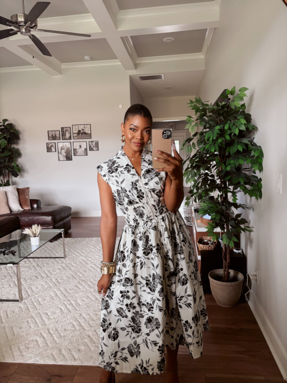 Woman wears the White and Black Floral Print Smocked Midi Dress-FINAL SALE and snaps a mirror selfie in a stylish room with plants.