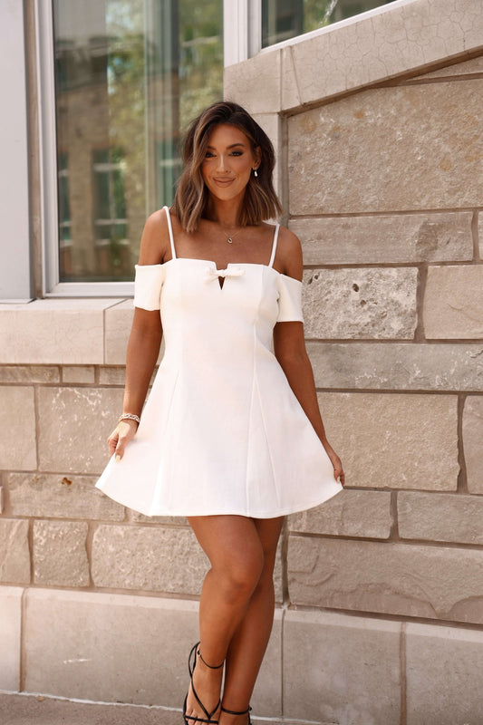 Woman in a White Cold Shoulder Flare Mini Dress - FINAL SALE stands outdoors by a stone wall, smiling and holding her dress.
