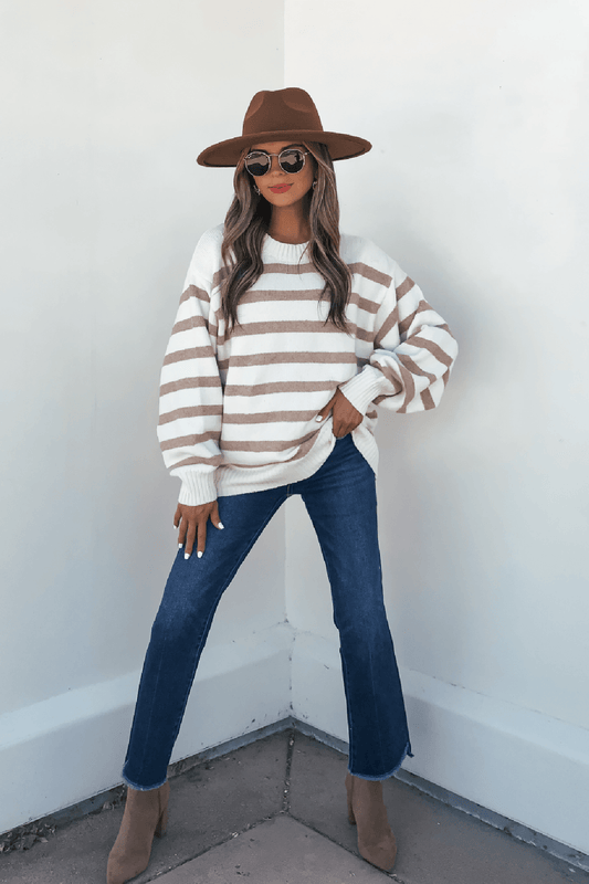 Woman in the Willow Cream Stripe Ribbed Sweater, jeans, sunglasses, boots, and brown hat poses by a white wall.