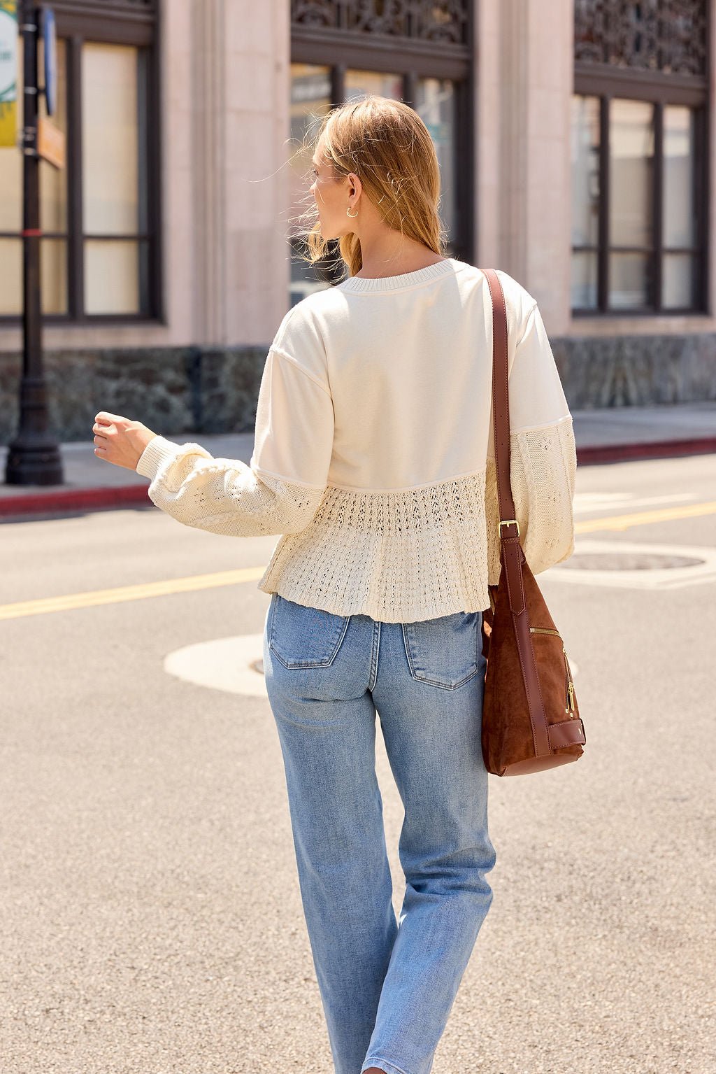 Someone in the Wren Textured Sweater - Cream and blue jeans walks on a sunny street, carrying a brown shoulder bag.