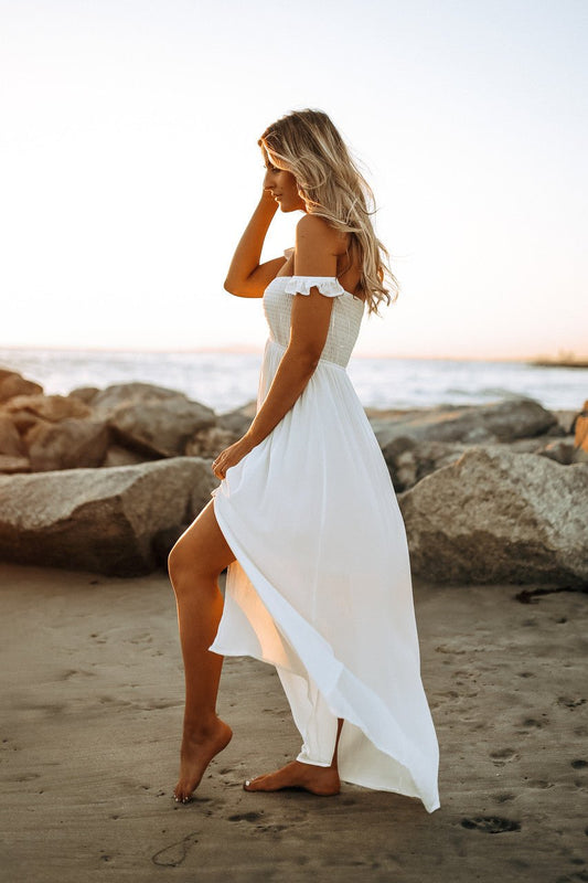 Barefoot woman in the Odella Off Shoulder Dress: Smocked Maxi in Off White walks on a sandy beach at sunset near rocks.