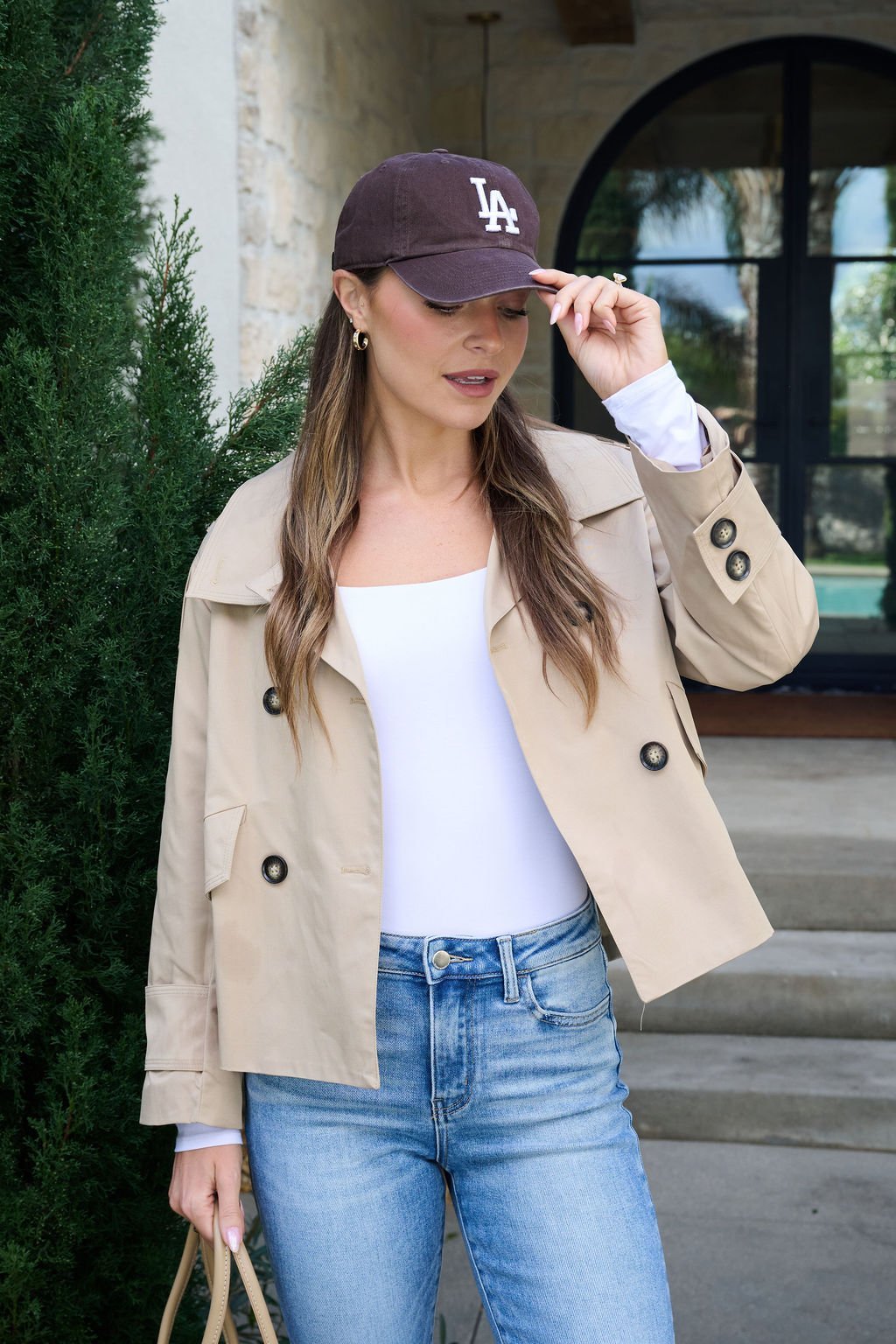 A woman in a tan jacket, white top, and blue jeans stands outdoors, holding a beige bag and adjusting her brown LA baseball cap, with greenery and a stone building in the background.