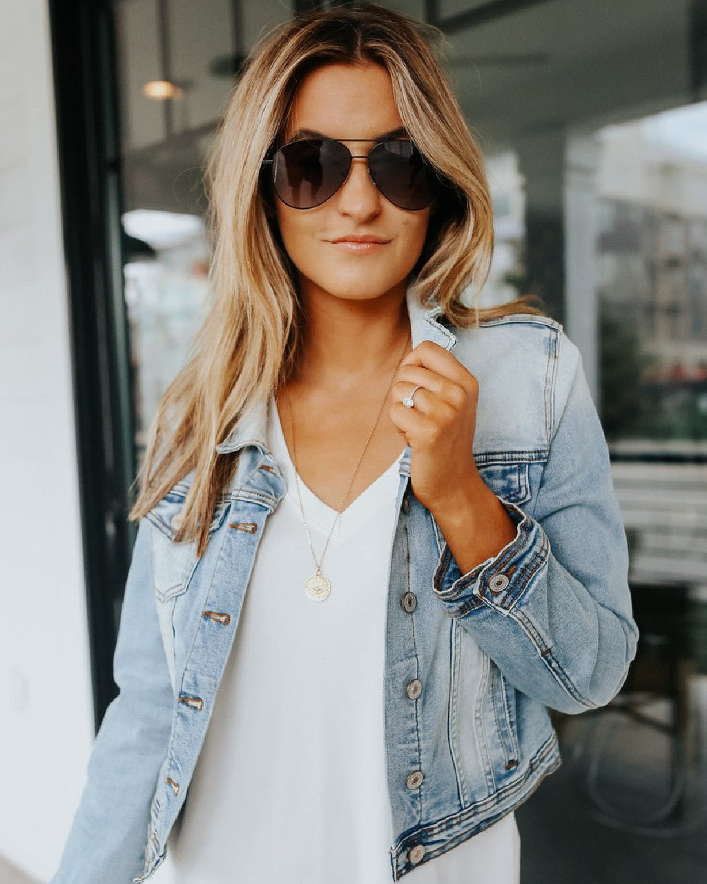 A woman wearing a light-wash blue denim jacket over a white top, accessorized with dark sunglasses, a delicate gold necklace, and a ring, standing in front of a modern glass storefront.