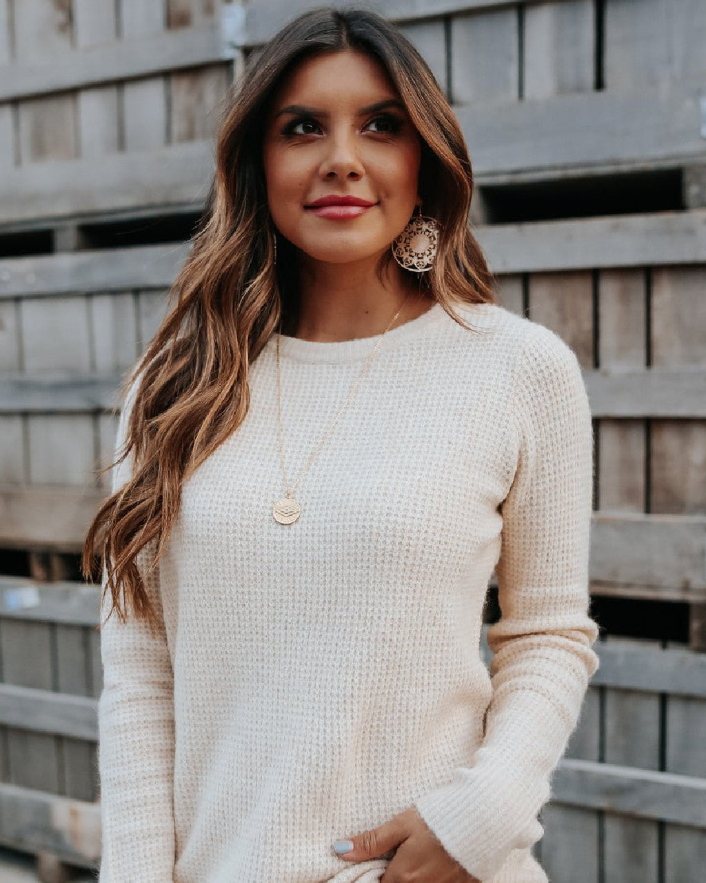 A woman with long wavy brown hair wearing a cream-colored waffle-knit long-sleeve sweater, standing in front of a rustic wooden fence.