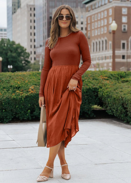 A woman wearing a rust-colored long-sleeve midi dress with a pleated skirt, standing outdoors in front of city buildings and hedges. She is holding a beige handbag and wearing beige heels.