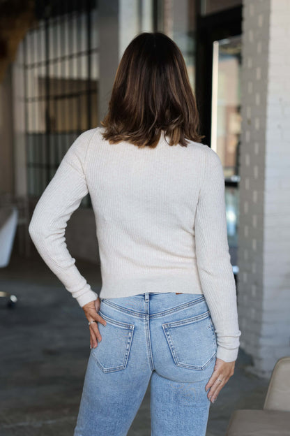 A woman stands indoors, facing away, wearing the Free People Oatmeal Ever After Pullover and light blue jeans.
