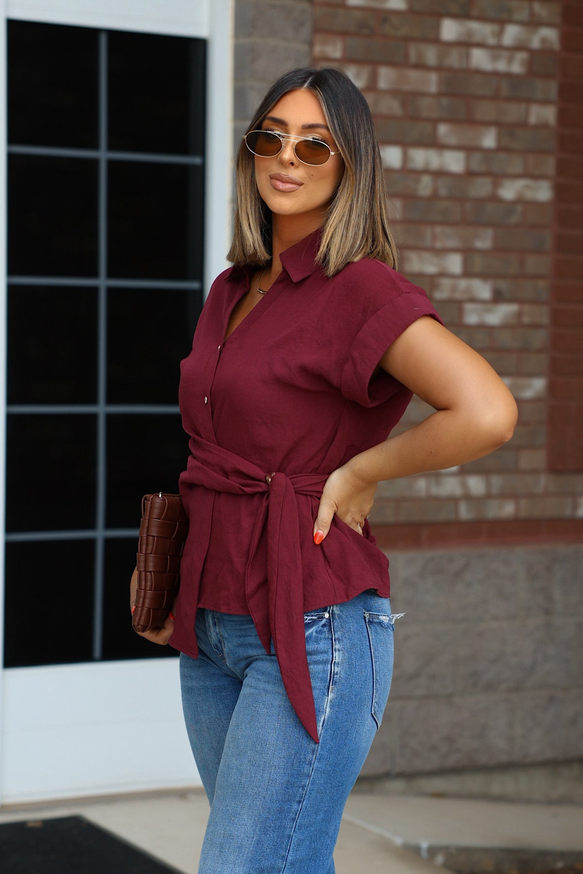 Woman in sunglasses wears the Burgundy Side Tie Buttoned Top with blue jeans and holds a brown clutch outside.