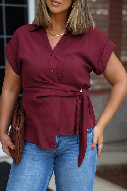 Woman in a Burgundy Side Tie Buttoned Top, blue jeans, holding a brown woven clutch outdoors.