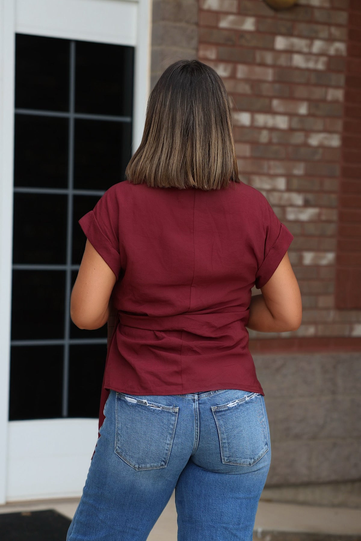 Someone in a Burgundy Side Tie Buttoned Top and ripped blue jeans stands facing away near a building entrance.