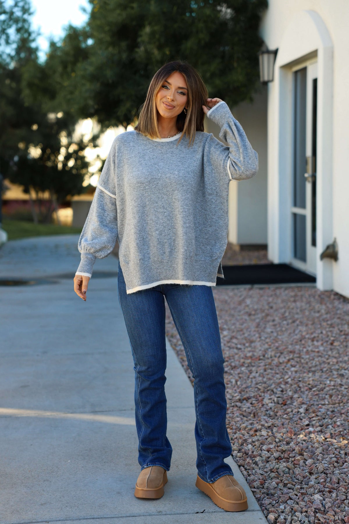 A woman outdoors smiles, touching her hair, wearing jeans, tan shoes, and a Grey Contrast Detail Pullover Sweater.