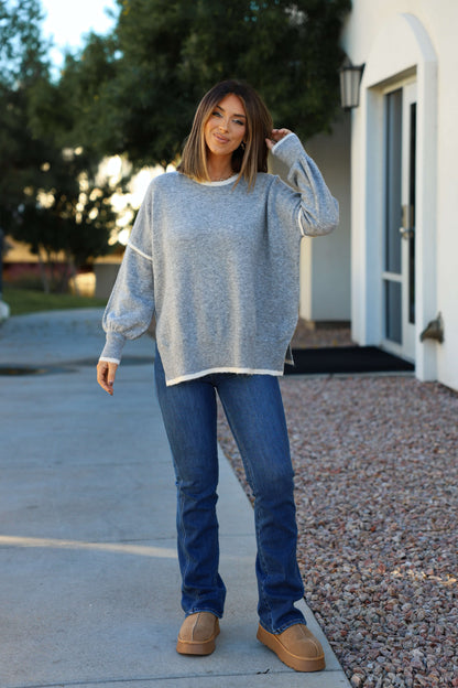 A woman outdoors smiles, touching her hair, wearing jeans, tan shoes, and a Grey Contrast Detail Pullover Sweater.