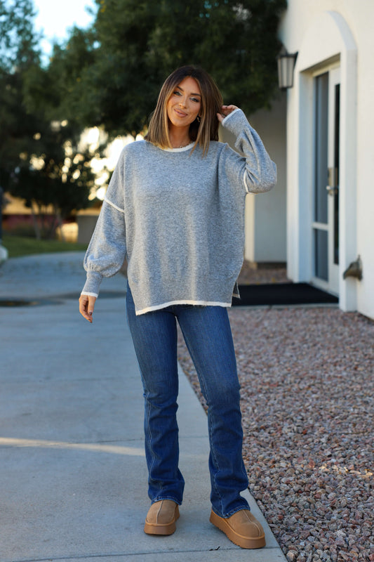 A woman outdoors smiles, touching her hair, wearing jeans, tan shoes, and a Grey Contrast Detail Pullover Sweater.