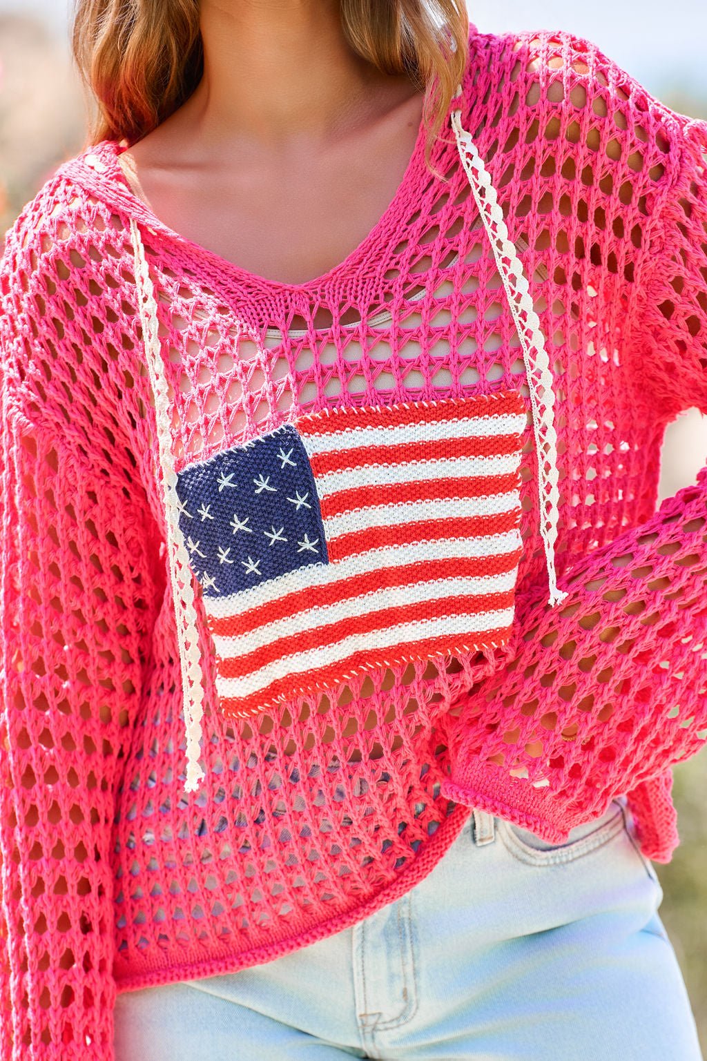 Woman outside in sunlight, wearing the All American Open Knit Hoodie in pink with an American flag patch and light blue jeans.