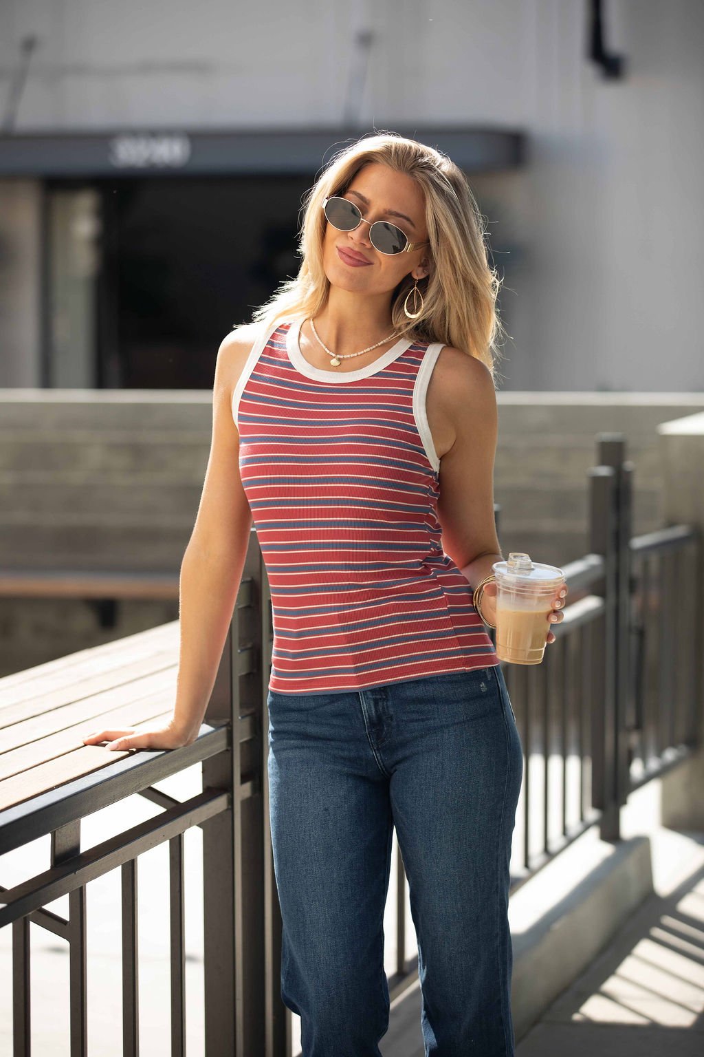 Woman in sunglasses wears the All-Day Stripe Tank Top - Red-FINAL SALE with jeans, holding a drink by an outdoor railing.