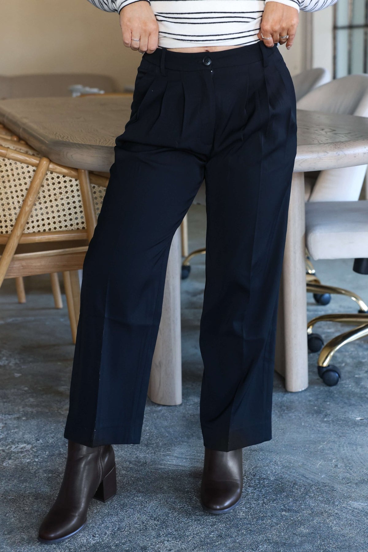 Person wears Anabelle Black Pleated Trousers, brown ankle boots, and a striped top while standing indoors by a table and chairs.