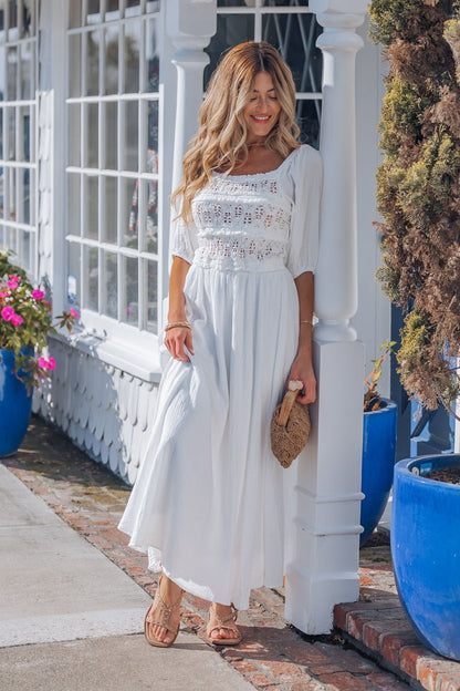 Woman wears the Angelica White Ruched Midi Dress, standing by a white building with blue flower pots and a woven clutch.