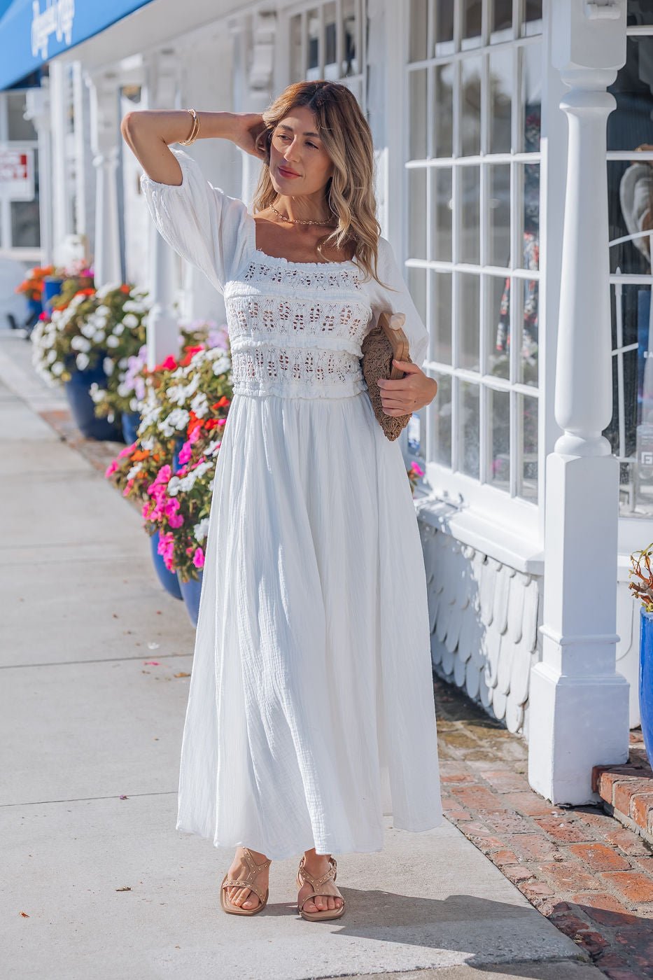 Woman wears the Angelica White Ruched Midi Dress - FINAL SALE, standing by a storefront with flowers, holding a clutch.