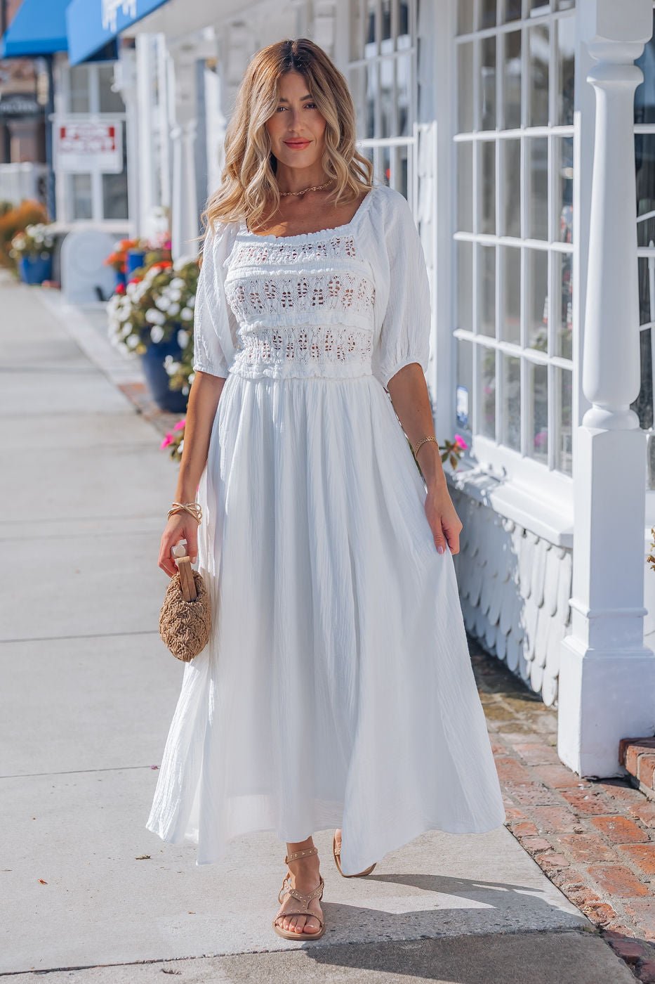 Woman walking in the Angelica White Ruched Midi Dress - FINAL SALE, paired with sandals and a woven bag on a sunny sidewalk.