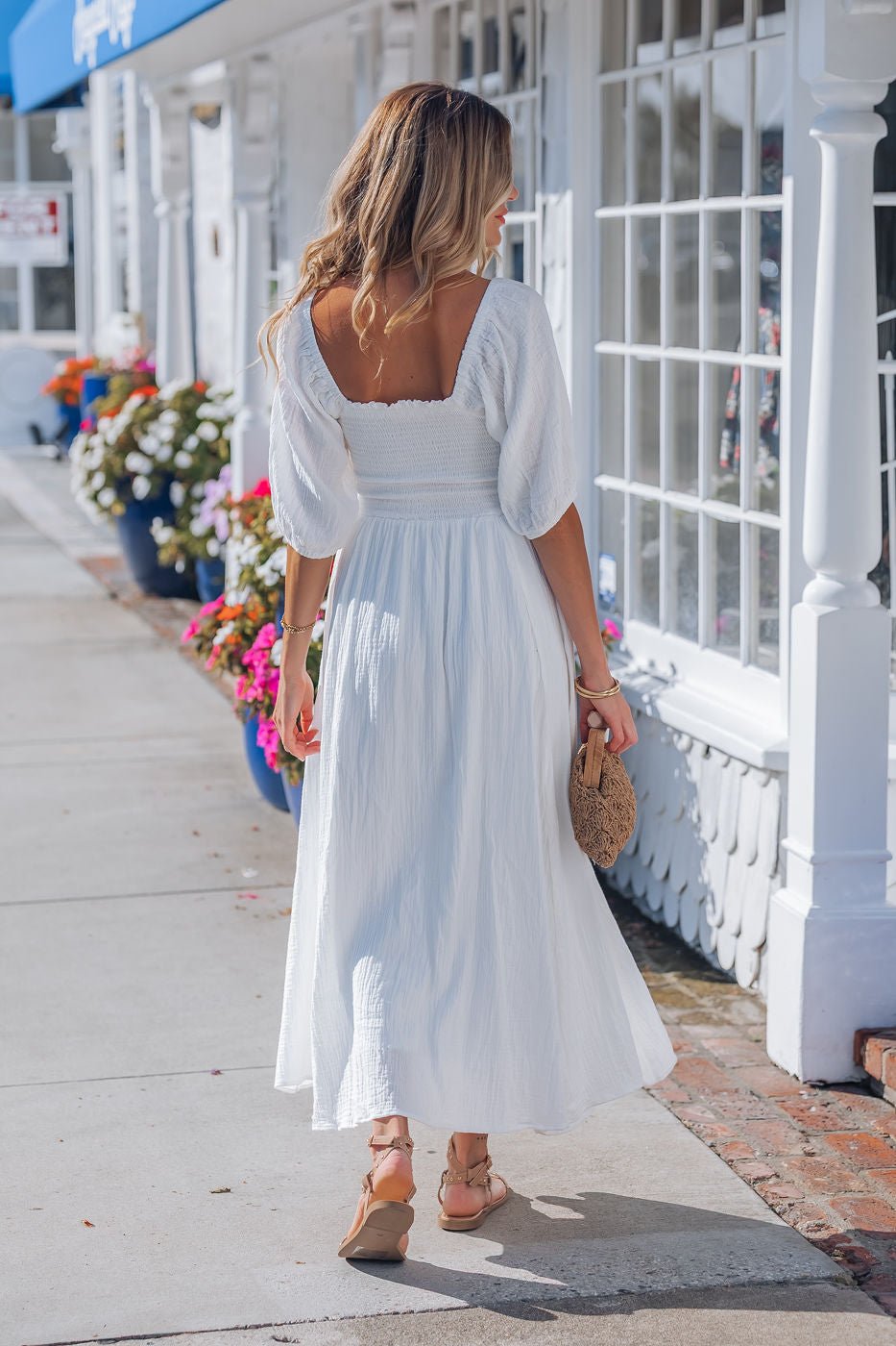 A woman in the Angelica White Ruched Midi Dress strolls on a sunny, flower-lined sidewalk with sandals and a small woven bag.