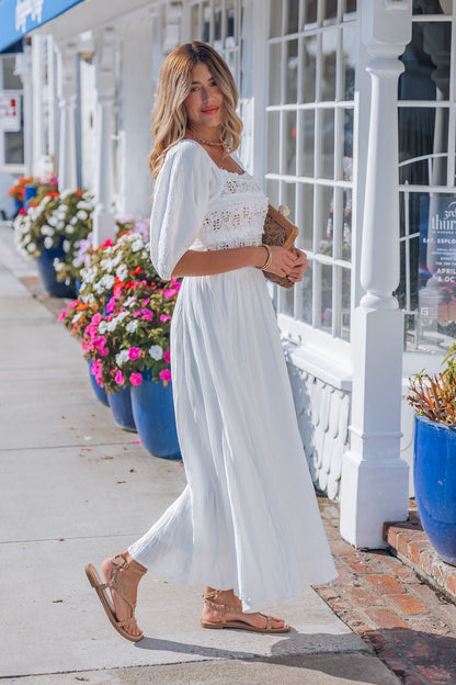Woman wearing the Angelica White Ruched Midi Dress - FINAL SALE stands on a sidewalk, holding a clutch and smiling.