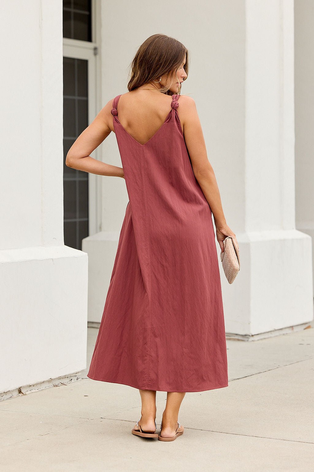 Woman in the Audrey Knot Strap Midi Dress - Burgundy stands outdoors, facing away, holding a beige clutch and wearing sandals.