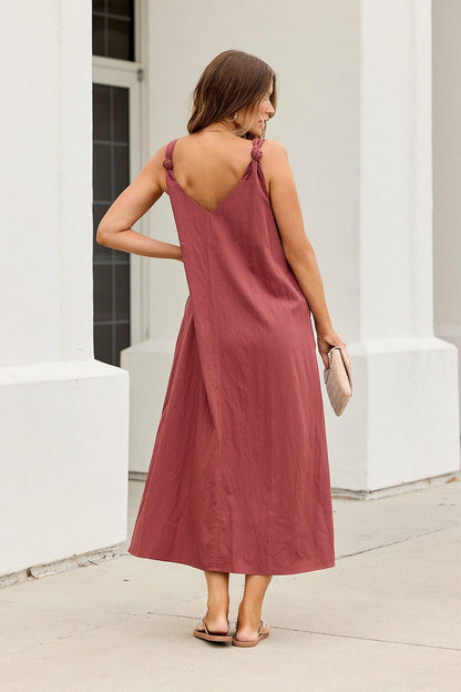Woman in the Audrey Knot Strap Midi Dress - Burgundy stands outdoors, facing away, holding a beige clutch and wearing sandals.