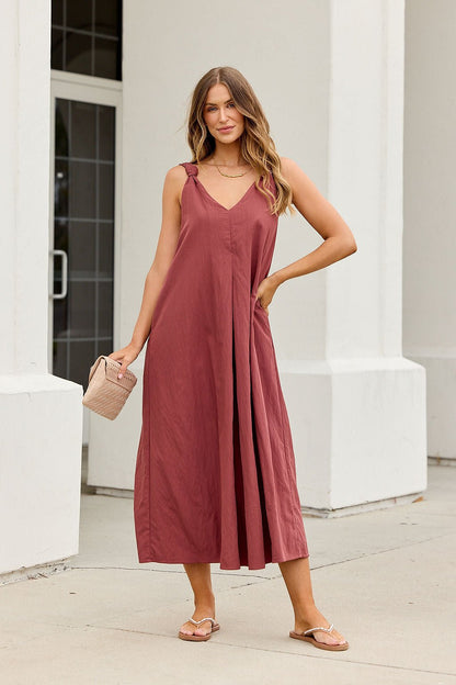 Woman in the Audrey Knot Strap Midi Dress - Burgundy stands outdoors with a purse near a white building.