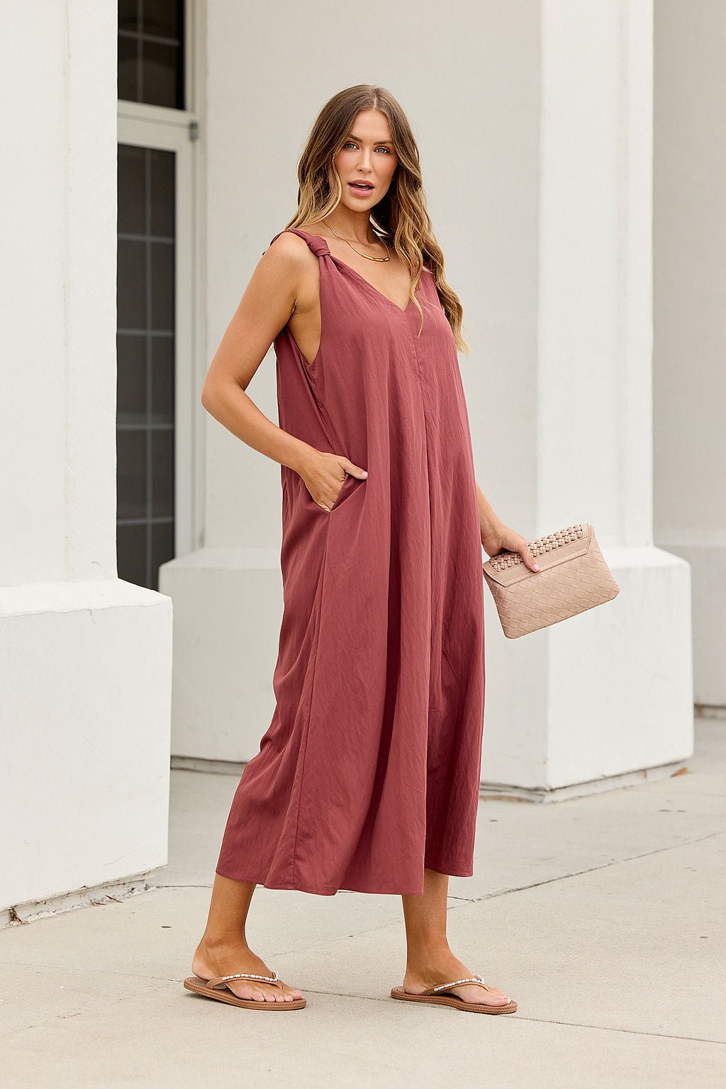 Woman wears the Audrey Knot Strap Midi Dress in burgundy with pockets, holding a beige clutch and standing on the sidewalk in sandals.